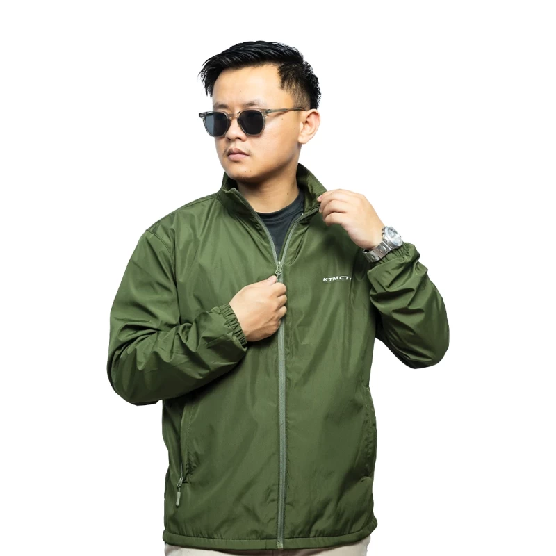A male model wearing sunglasses looks off to the side while adjusting the collar of his olive green KTM CTY windbreaker, windcheater. The lightweight windguard jacket features a full-zip front, elastic cuffs, and a small white logo on the chest